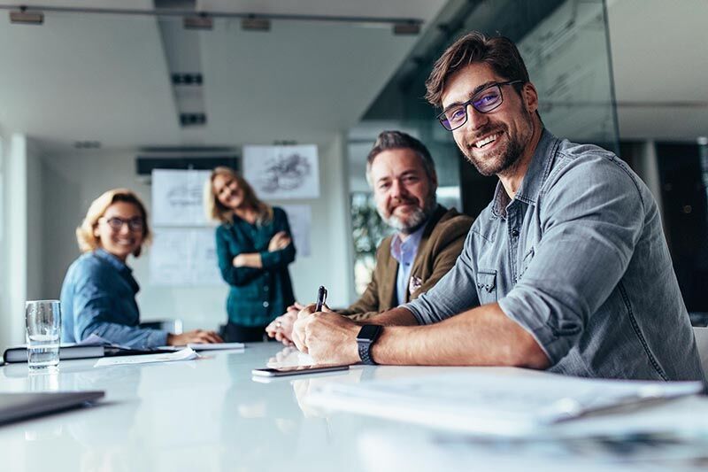 Eine Gruppe Menschen sitzt in einem Büro. Ein Mann mit Brille und Stift in der Hand im Vordergrund.